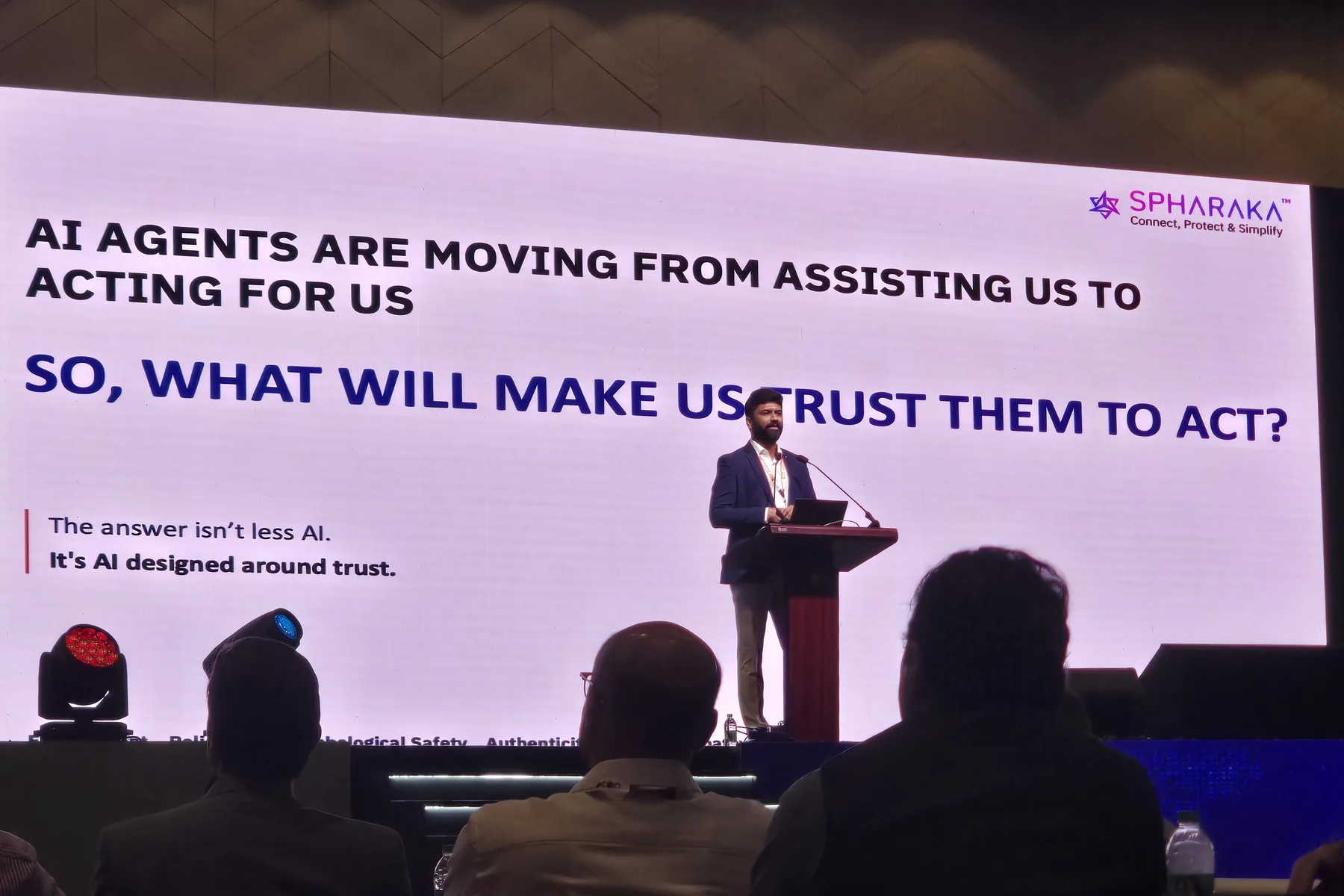 Rajeev Raj speaking at a lectern at C0C0N 2026, beside a screen reading that AI agents are moving from assisting us to acting for us, and asking what will make us trust them to act.
